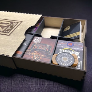 May include: A wooden box with a lid that says "20 Strong" on the front. The box is open and contains a variety of game components, including cards, a rulebook, and a dial. The box is designed to store and organize the components of a board game.
