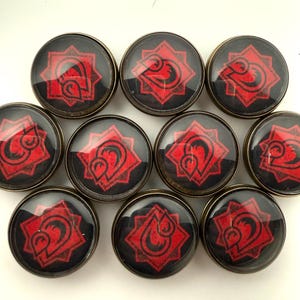 May include: A collection of nine circular buttons featuring a black background with a red, eight-pointed star design containing a stylized graphic symbol. Each button is encased in a clear, domed glass cover and framed by a metallic bronze-toned rim.
