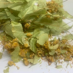 May include: A collection of dried linden flowers and light green leaves. The botanical material features small, shrivelled yellow blossoms mixed with thin, papery, heart-shaped foliage. The dried herbs are scattered across a plain white surface.
