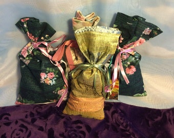 May include: Three fabric sachets with floral patterns. The sachets are tied with pink and white ribbon. The sachets are on a purple background.
