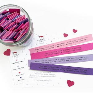 May include: A clear glass jar filled with pink and purple date night ideas. Several strips of paper with date night suggestions are displayed, along with heart-shaped cutouts. Text on the strips includes ideas like "Enjoy wine" and "Turn your workout into a date."