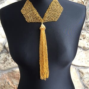 May include: A gold beaded necklace with a bib-style collar and a long tassel pendant. The necklace is displayed on a black mannequin against a stone wall background. The intricate beadwork creates a textured, shimmering effect.