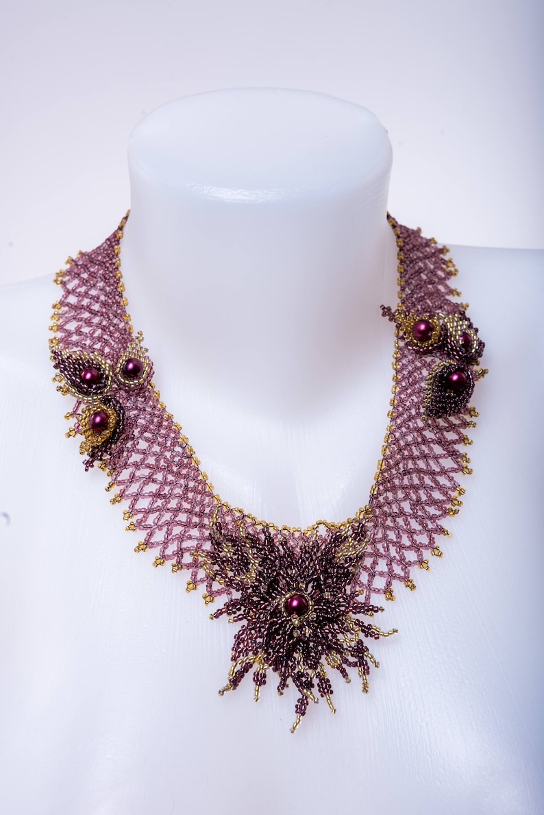 May include: A delicate beaded necklace with a floral design. The necklace is made of small, purple and gold beads, and features a large, purple flower in the center.