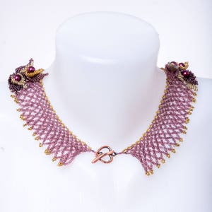 May include: A delicate beaded necklace with a copper clasp. The necklace is made of small, purple and gold beads woven into a intricate pattern.