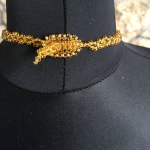 May include: A golden beaded choker necklace. The necklace is made of small, round, golden beads strung together to form a delicate, yet intricate design. The clasp is a larger cluster of beads, adding a focal point to the necklace.
