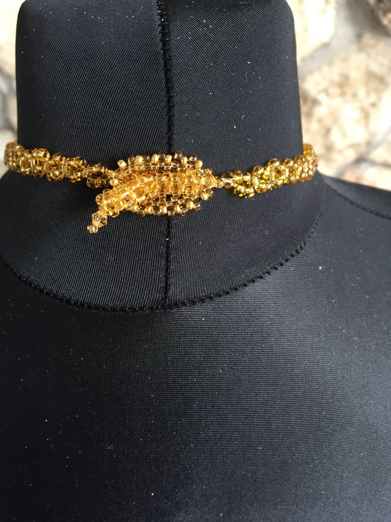 May include: A golden beaded choker necklace. The necklace is made of small, round, golden beads strung together to form a delicate, yet intricate design. The clasp is a larger cluster of beads, adding a focal point to the necklace.
