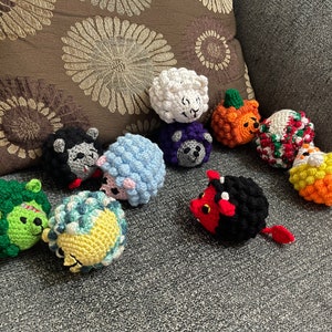 May include: A collection of 11 crocheted stuffed toys, including a sheep, a pumpkin, a snowman, a green monster, a blue and yellow creature, a black and red devil, a black sheep, a purple creature, a blue sheep, and two orange and yellow creatures.