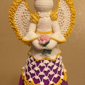 May include: A crocheted angel figurine with white and yellow yarn, holding a pink rose. The angel is wearing a white dress with yellow trim and a yellow halo. The angel is standing on a purple base.