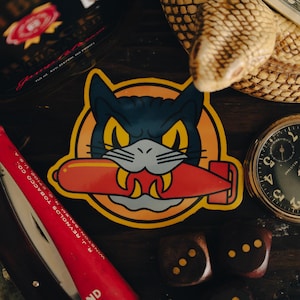 May include: A sticker featuring a cartoon cat with a navy blue face, yellow eyes, and a red torpedo in its mouth. The sticker is set against an orange circle. Other items include a pocket watch, dice, and a red tobacco product.