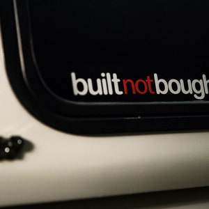 Könnte beinhalten: Ein weißer Aufkleber mit dem Text "built not bought" in roten und weißen Buchstaben auf schwarzem Hintergrund.
