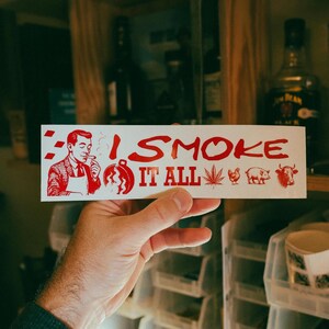 May include: A white sticker with red text and illustrations. The text reads "I SMOKE IT ALL" with images of a man smoking, a grill, a cannabis leaf, a chicken, a pig, and a cow.