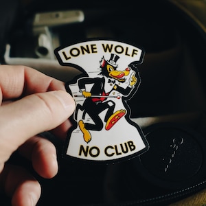 Puede incluir: Una pegatina con un lobo de dibujos animados con esmoquin, sombrero de copa y pajarita, corriendo con un bastón. La pegatina tiene las palabras "LONE WOLF" encima del lobo y "NO CLUB" debajo. El lobo es amarillo, negro y rojo.