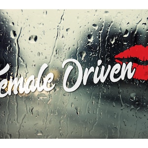 Puede incluir: Calcomanía de vinilo blanco con el texto "Female Driven" en una fuente cursiva. Una impresión de labios rojos está en el lado derecho de la calcomanía. La calcomanía está en una ventana de automóvil con gotas de lluvia.