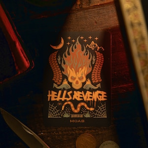 May include: A black and orange sticker with a skull and flames. The sticker says "Hells Revenge" and "MOAB" in a stylized font.