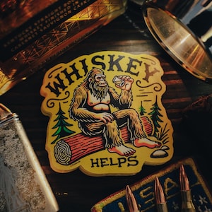 May include: A yellow sticker with a cartoon illustration of a Bigfoot sitting on a log and drinking from a glass. The text "Whiskey Helps" is printed below the Bigfoot.