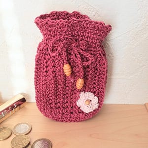 May include: A handmade, magenta-colored crochet drawstring pouch with a scalloped top edge. The pouch features a small, light pink crocheted flower and two wooden beads. Makeup and coins are visible on the wooden surface.