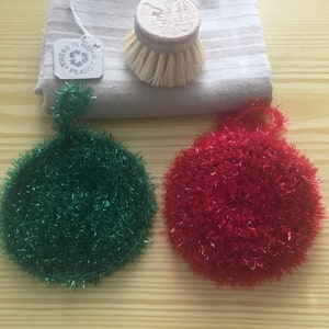 Peut inclure: Deux éponges rondes, verte et rouge, en fil bouclé. Les éponges sont sur une surface en bois. Il y a une étiquette qui dit "Il n'y a pas de planète B".