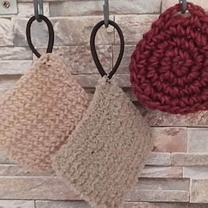 May include: Two square-shaped beige crochet dishcloths and one circular dark red crochet dishcloth, each featuring a black elastic loop for hanging. The items are crafted with a textured yarn pattern and are displayed suspended from metal hooks.