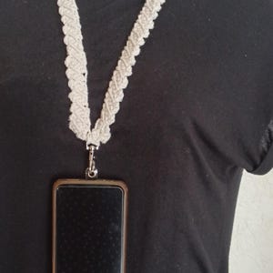 Peut inclure: Un cordon de macramé crème avec un fermoir argenté tenant un smartphone. Le cordon est fait d'un matériau épais et tissé et se porte autour du cou. Le téléphone est dans un étui marron.