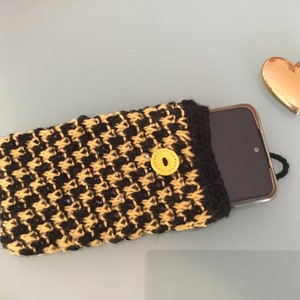 Peut inclure: Une coque de téléphone en crochet noir et jaune avec un bouton jaune. La coque est posée sur une surface bleu clair avec un objet en forme de cœur doré à droite.