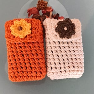 Puede incluir: Dos fundas de teléfono de ganchillo hechas a mano. Una es naranja con una flor amarilla, la otra es blanquecina con una flor marrón. Ambas tienen un cierre de solapa y un patrón de puntada texturizada. Las fundas están diseñadas para guardar un teléfono.