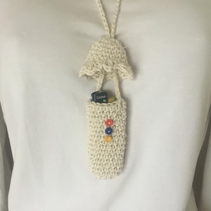 Peut inclure: Une pochette blanche en crochet avec un dessus en crochet et trois boutons colorés. La pochette est suspendue à un cordon blanc autour du cou d'une personne. La pochette contient un petit objet bleu et jaune.