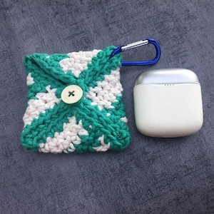 May include: A handmade crochet case in green and white, secured with a wooden button. A blue carabiner clip is attached. Beside it is a white and silver wireless earbud charging case.