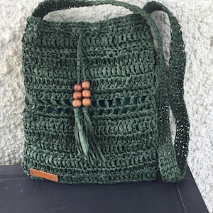 May include: A dark green woven shoulder bag with a drawstring closure and a long strap. The bag features a decorative tassel with wooden beads. A small brown leather tag is attached to the front.