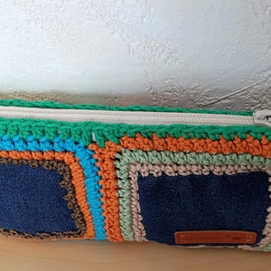 Può includere: Una pochette rettangolare con cerniera caratterizzata da due quadrati in denim blu scuro incorniciati da bordi all'uncinetto multicolore in verde, arancione, marrone e azzurro. Una zip beige corre lungo la parte superiore con una nappa.