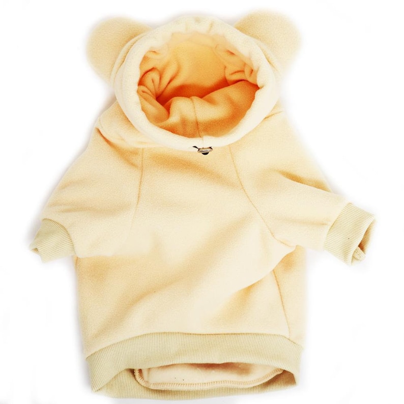 Pu&ograve; includere: Un maglione con cappuccio in pile giallo chiaro per cani con orecchie da orso. Il maglione ha una cerniera e un orlo a coste.