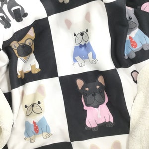 May include: A white and black checkered fleece blanket with a repeating pattern of French Bulldogs wearing different outfits. The dogs are wearing a variety of outfits, including a blue shirt with a red tie, a pink hoodie, and a white shirt with a blue collar.