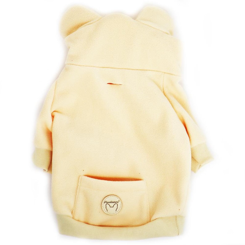 Pu&ograve; includere: Una felpa con cappuccio gialla in pile per cani con cappuccio e una tasca sul retro. La felpa ha un piccolo patch in pelle marrone con la scritta "Pawsitive" e un'impronta di zampa.