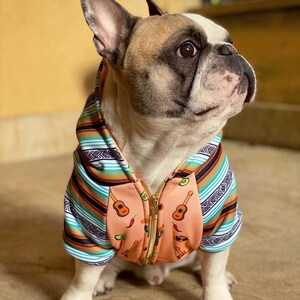 May include: A French bulldog wearing a peach-colored hoodie with a colorful striped pattern and a zipper. The hoodie has a pattern of guitars, chili peppers, and cacti.