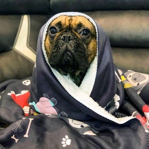 May include: A French bulldog puppy is snuggled in a black and white fleece blanket with a cartoon dog print. The blanket has a white sherpa lining.