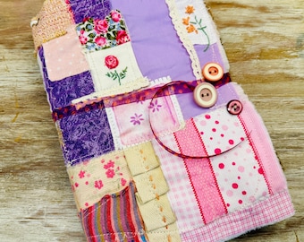 May include: A fabric-covered journal featuring a patchwork design of purple, pink, and white textiles. The cover includes floral embroidery, lace trim, a thin ribbon tie, and three round buttons in shades of orange, cream, and dark pink.