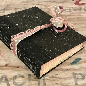 May include: A black journal with white paint splatter and a pink and polka dot ribbon closure. The edges of the pages are visible, and the journal is on a wooden surface.