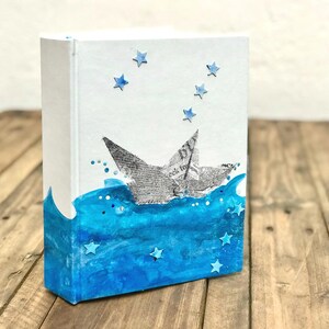 May include: A white, rectangular decorative box with a nautical theme. The box features a paper boat design, blue water, and star accents. The boat is made from newspaper print, and the water is painted in shades of blue.
