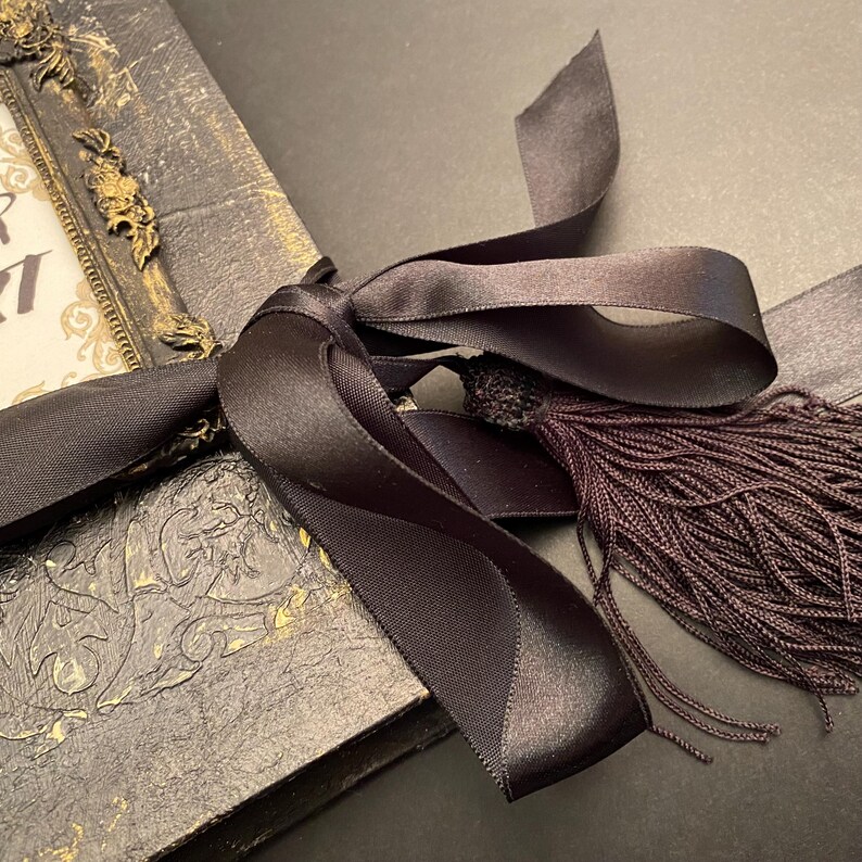 May include: A black leather-bound journal with gold accents and a black satin ribbon tied around it with a tassel.