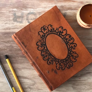 May include: A brown leather journal with a decorative black oval frame design on the cover. The journal is closed and lies on a wooden surface, with a candle and pencils nearby.