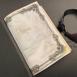 May include: An antique book with a black cover and a lace-trimmed page. The page is decorated with a floral design and has the word "Signed" written on it.