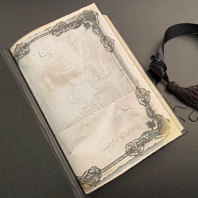 May include: An antique book with a black cover and a lace-trimmed page. The page is decorated with a floral design and has the word "Signed" written on it.