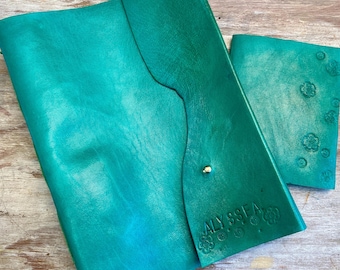 Puede incluir: Un diario de cuero turquesa y un pequeño cuaderno a juego. El diario tiene un cierre de solapa con un botón dorado. El diario está grabado con el nombre "ALYSSSA" y diseños florales. El cuaderno tiene diseños florales en relieve.
