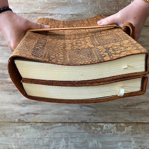 May include: Two brown leather-bound journals with a decorative pattern on the cover. The journals are closed and stacked on top of each other.