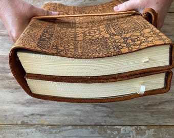 May include: A tan leather journal cover featuring a dark brown intricate lace pattern embossed on the surface. The cover wraps around two thick books with cream-coloured pages and includes a leather strap closure and small white ribbon bookmarks.