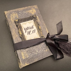May include: A black and gold journal with a decorative frame and a black ribbon tied around it. The frame has the text "YOUR TEXT" inside.