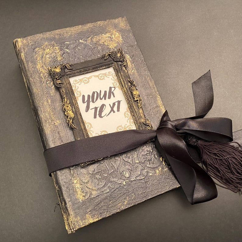 May include: A black and gold journal with a decorative frame and a black ribbon tied around it. The frame has the text "YOUR TEXT" inside.
