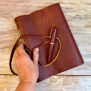 May include: A burgundy leather journal with a matching pen. The journal is closed with a leather strap. The pen has a gold-colored clip. The journal and pen are on a light-colored wooden surface.