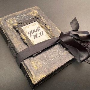 May include: A black and gold journal with a decorative frame and a black ribbon tied around it. The frame has the text "your text" inside.