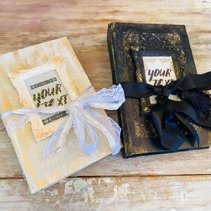May include: Two decorative journals with a distressed, vintage look. One journal is white with a lace ribbon and a frame that says "Your Text". The other journal is black with a black satin ribbon and a frame that says "Your Text".