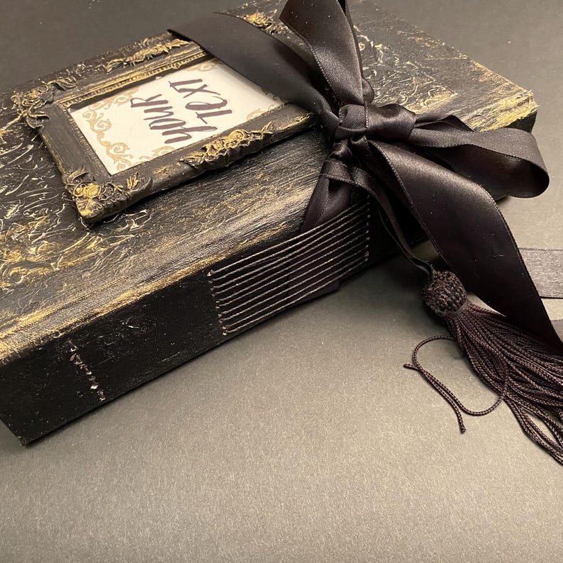 May include: A black book with a gold-painted cover and a black ribbon tied around it. The book has a small frame on the front with the words "Your text" written inside. The book is sitting on a gray surface.